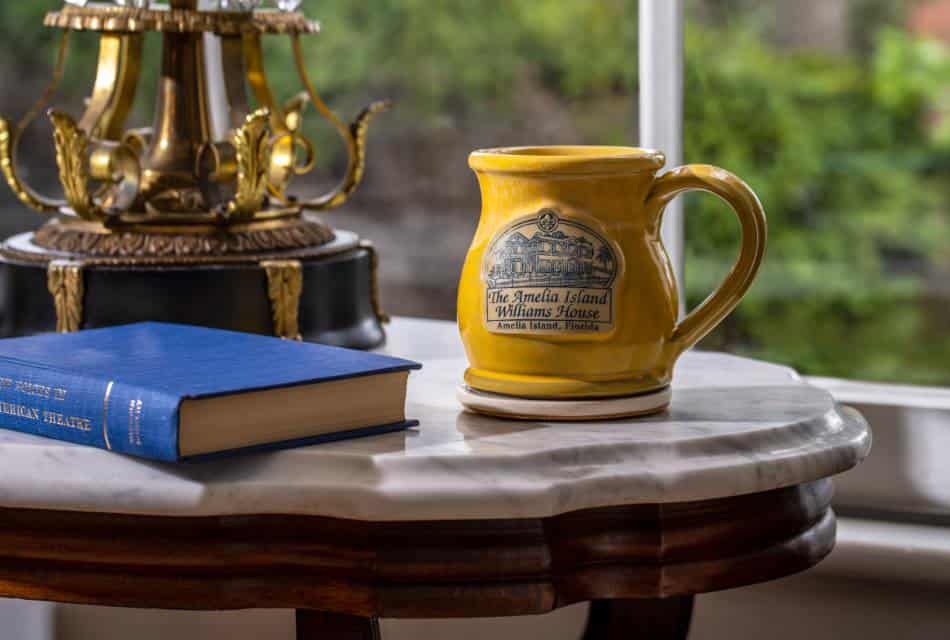 Close up view of yellow stoneware mug with The Amelia Island Williams House logo and blue book sitting on wooden stool with marble top