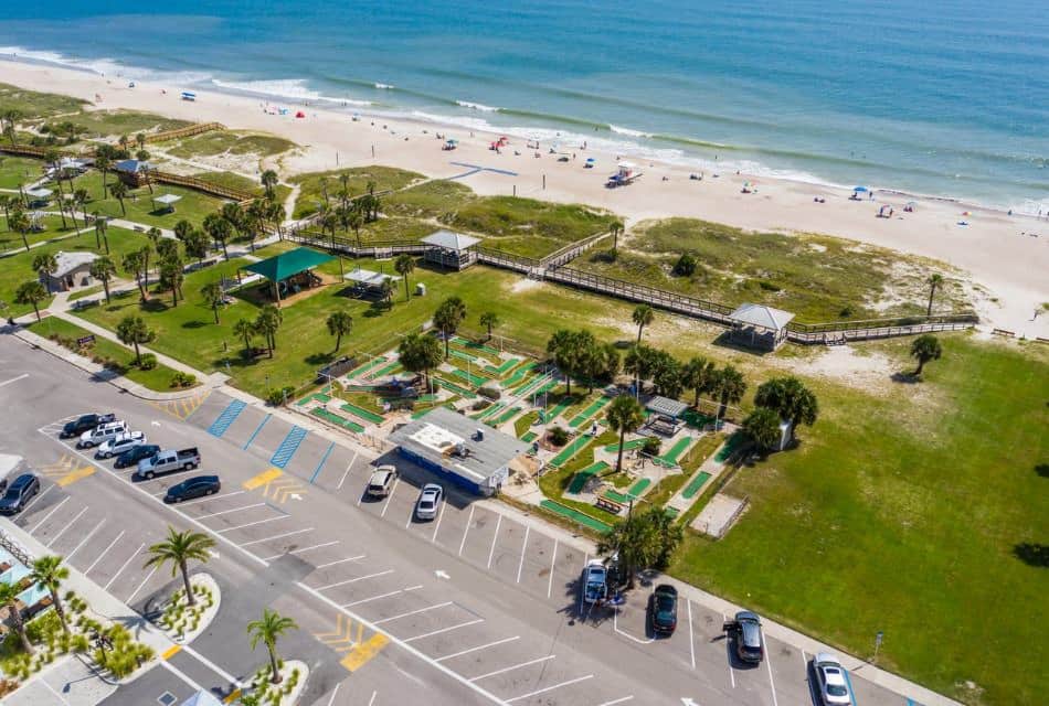 Aerial view of beach park with a parking lot, mini golf course, green grass, and access to the beach