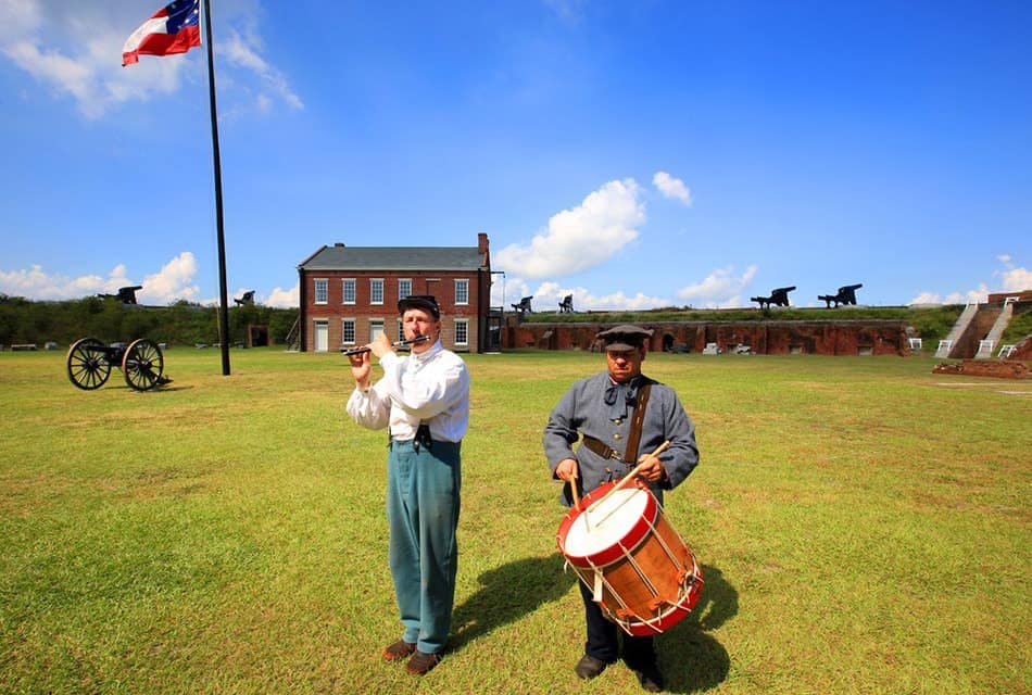 Two people standing near an old fort dressed up in old colony clothes with one playing a flute and another playing a drum