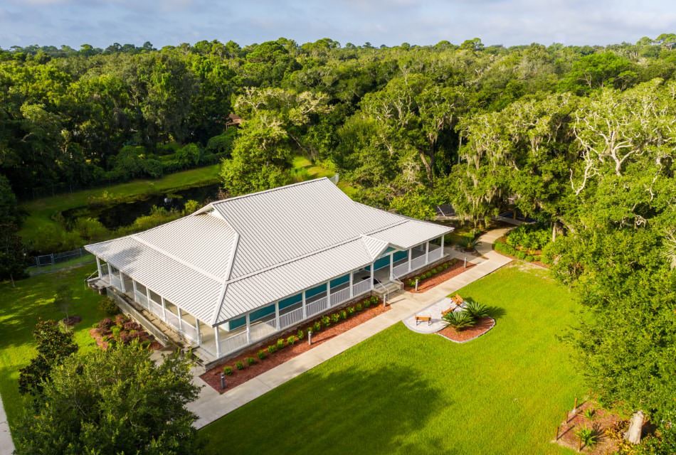 Exterior view of museum with white roof and white railing porch surrounded by green grass, shrubs, and large trees