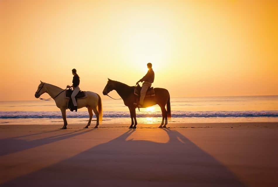 Two people on horseback standing on the sand near the water with the setting sun in the background