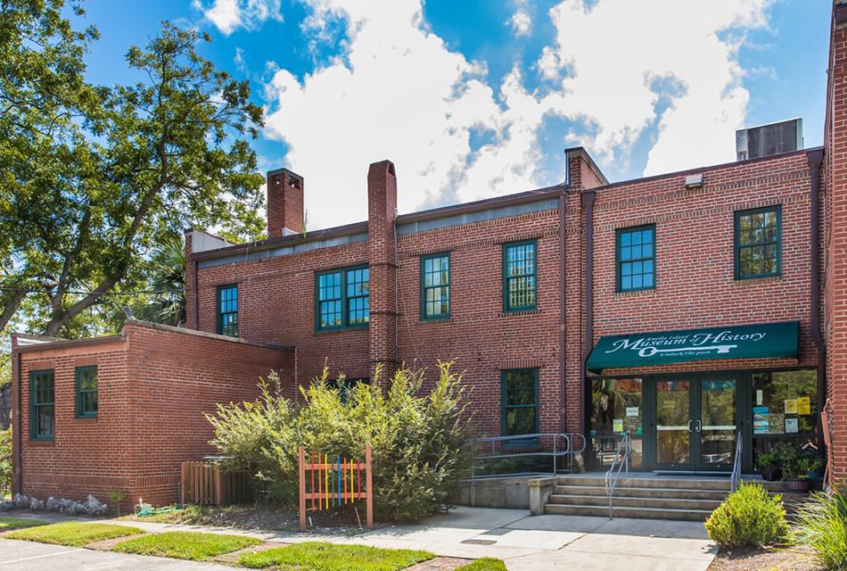 Exterior view of a red brick building with green trim surrounded by green bushes and trees