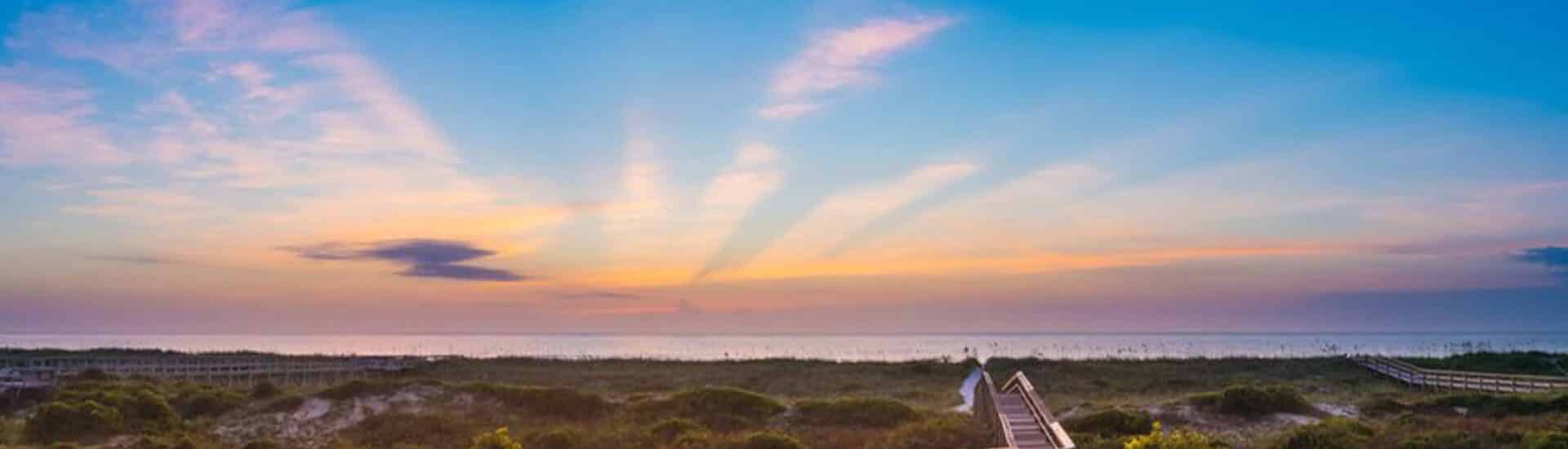 Long wooden walkway through green shrubs to the beach with water and setting sun in the background