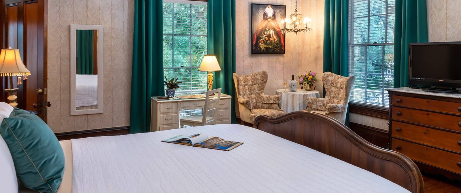 Bedroom with wallpaper, hardwood flooring, wooden bed, white bedding, white desk and chair, sitting area, and wooden dresser