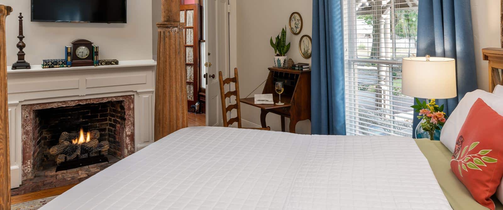 Bedroom with cream walls, hardwood flooring, bed with large wooden posts, white bedding, fireplace, and large windows