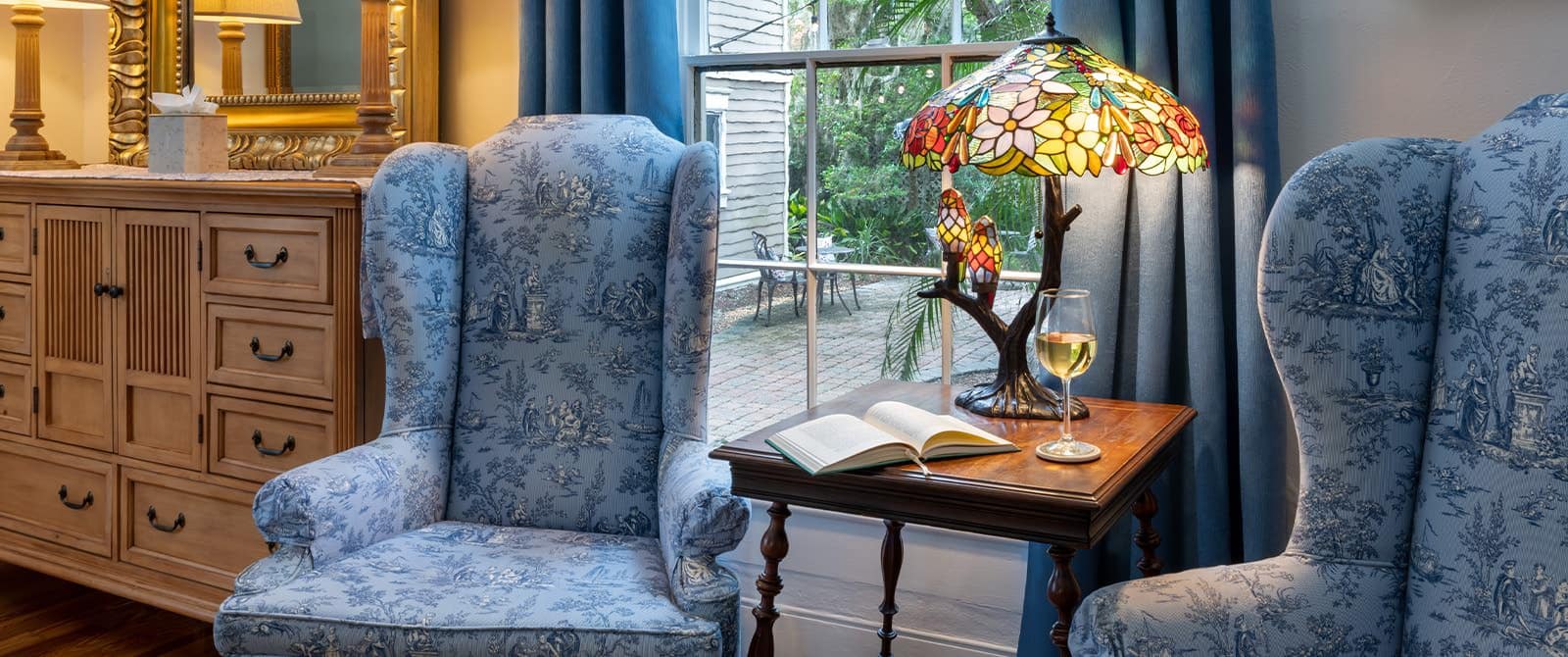 Close up view of a bedroom's sitting area with blue upholstered armchairs, wooden table with stained glass lamp, and light wooden dresser