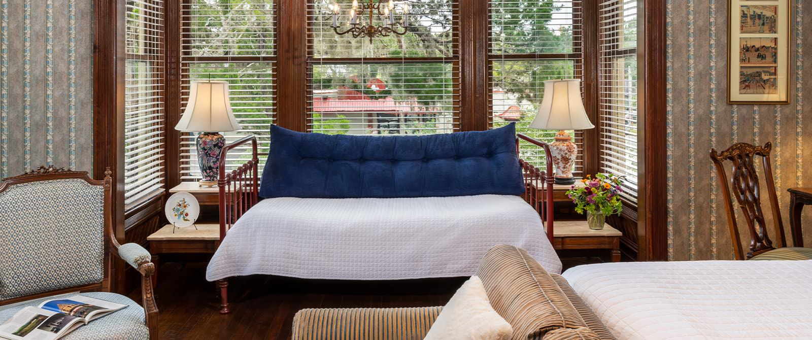 Bedroom with ornate wallpaper, hardwood flooring, daybed with metal frame, white bedding, upholstered loveseat, and dark wooden desk with chair