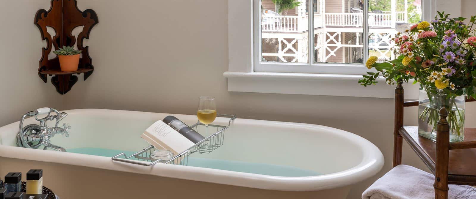 Bathroom with white walls, white clawfoot tub, wooden shelf with a vase of multicolored flowers, and window