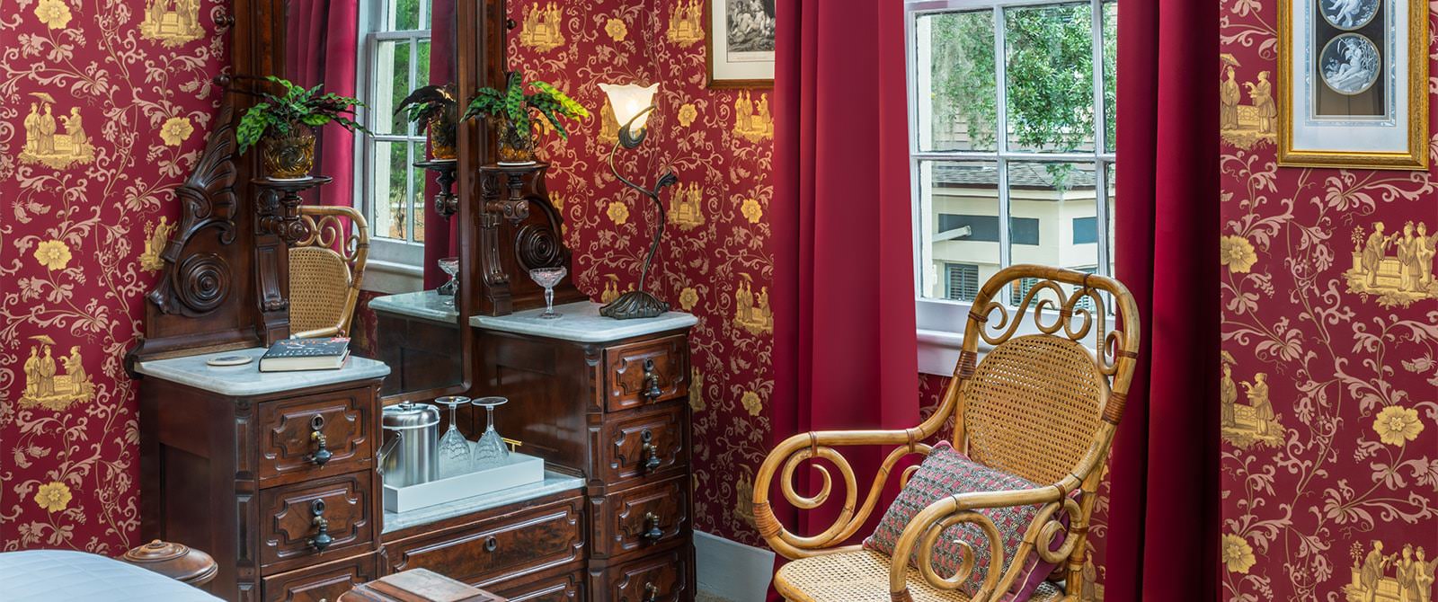 Bedroom with red ornate wallpaper, carpeting, antique wooden dresser with mirror, ornate wicker chair, and window