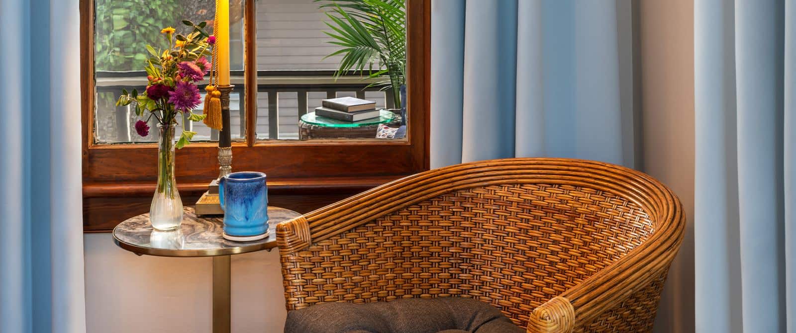 Close up view of light colored wicker chair with dark taupe cushion next to small table with blue mug, candle, and vase with flowers