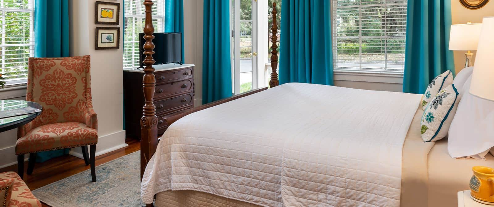 Bedroom with white walls, hardwood flooring, wooden four-poster bed, white bedding, wooden dresser, and sitting area