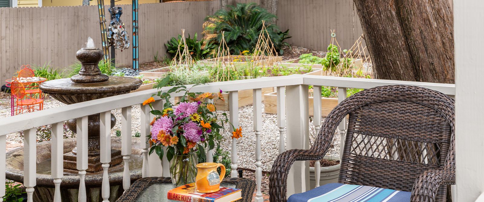 Close up view of wicker chair and table with orange mug, book, and vase of flowers with raised garden beds in the background