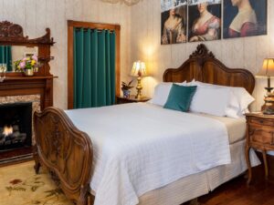 Bedroom with wallpaper, hardwood flooring, wooden bed, white bedding, and fireplace with wooden mantel and mirror