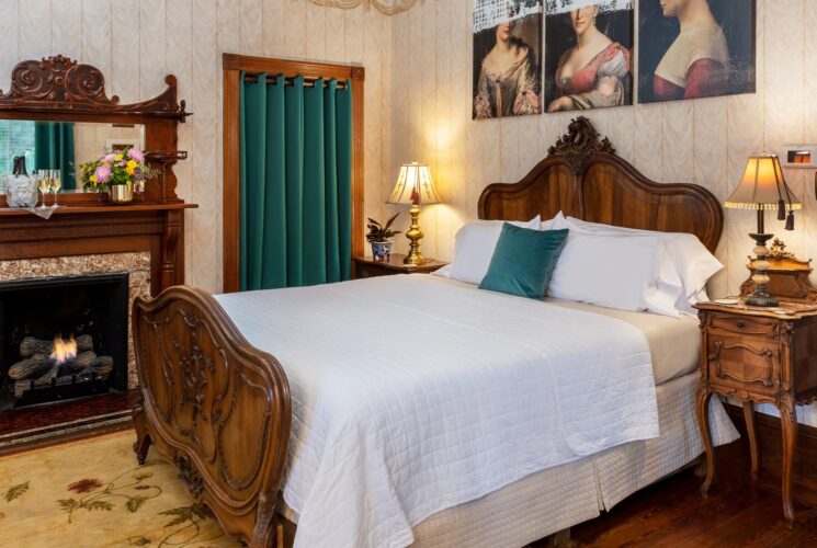 Bedroom with wallpaper, hardwood flooring, wooden bed, white bedding, and fireplace with wooden mantel and mirror