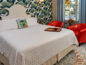 Bedroom with ornate wallpaper, hardwood flooring, white headboard, white bedding, and sitting area with red armchairs