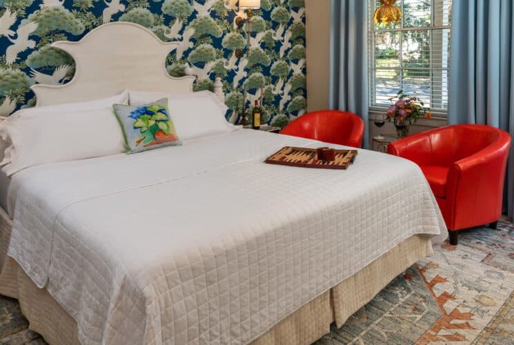 Bedroom with ornate wallpaper, hardwood flooring, white headboard, white bedding, and sitting area with red armchairs