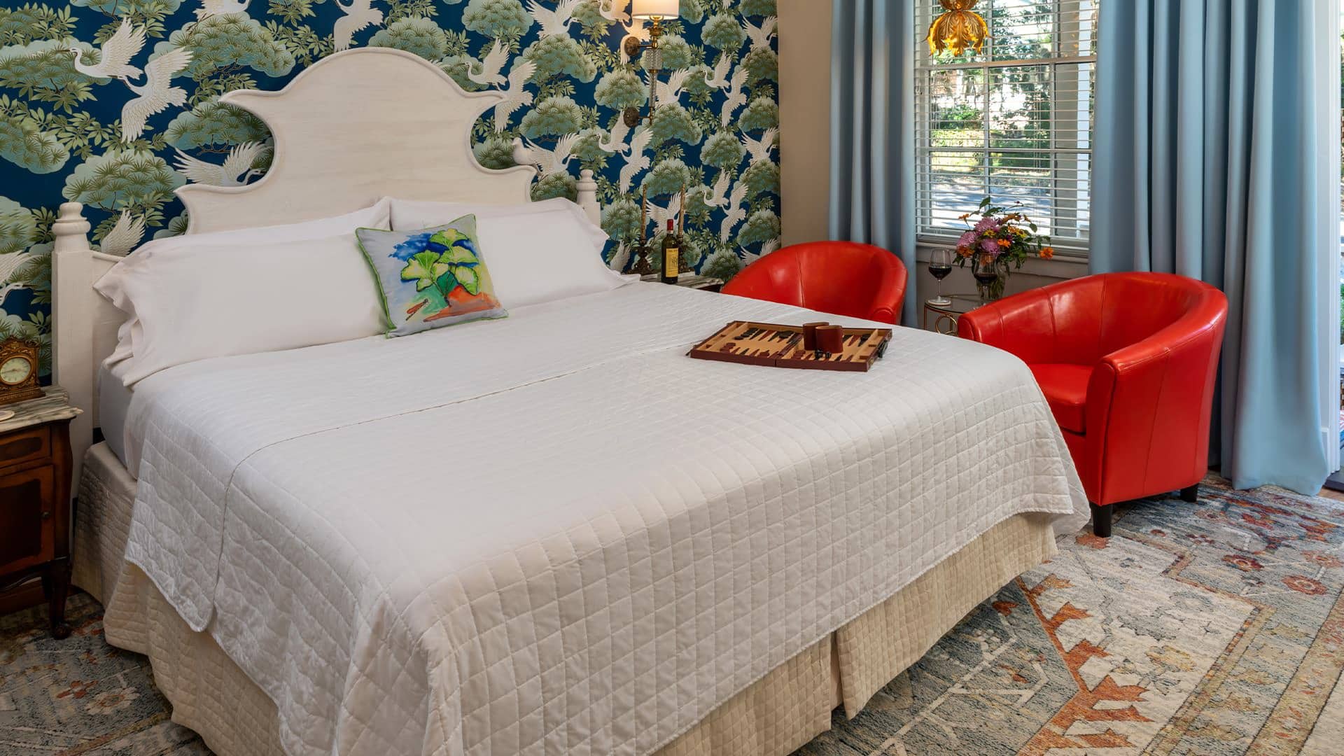 Bedroom with ornate wallpaper, hardwood flooring, white headboard, white bedding, and sitting area with red armchairs