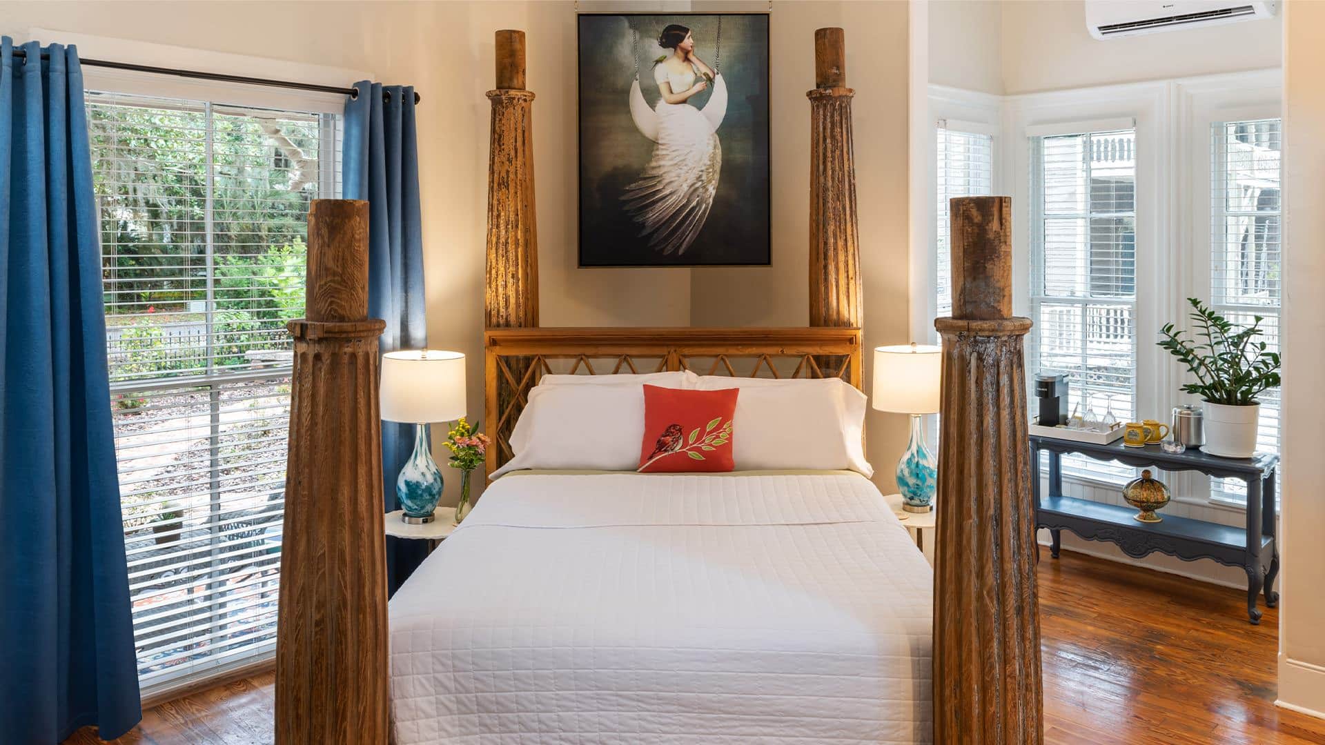 Bedroom with cream walls, hardwood flooring, bed with large wooden posts, white bedding, and large windows