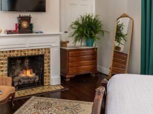 Bedroom with white walls, hardwood flooring, white bedding, fireplace, wooden dresser, and antique armchair