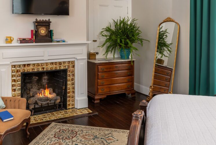 Bedroom with white walls, hardwood flooring, white bedding, fireplace, wooden dresser, and antique armchair