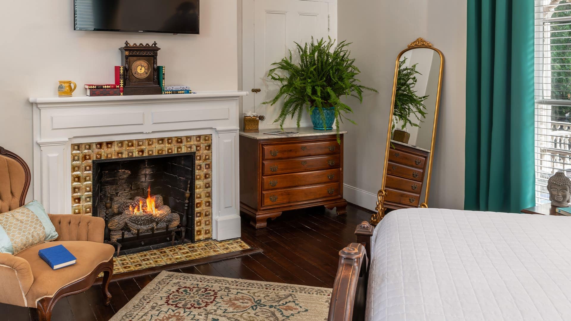 Bedroom with white walls, hardwood flooring, white bedding, fireplace, wooden dresser, and antique armchair