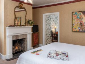 Bedroom with wallpaper, white bedding, fireplace with white mantel, and ornate mirror