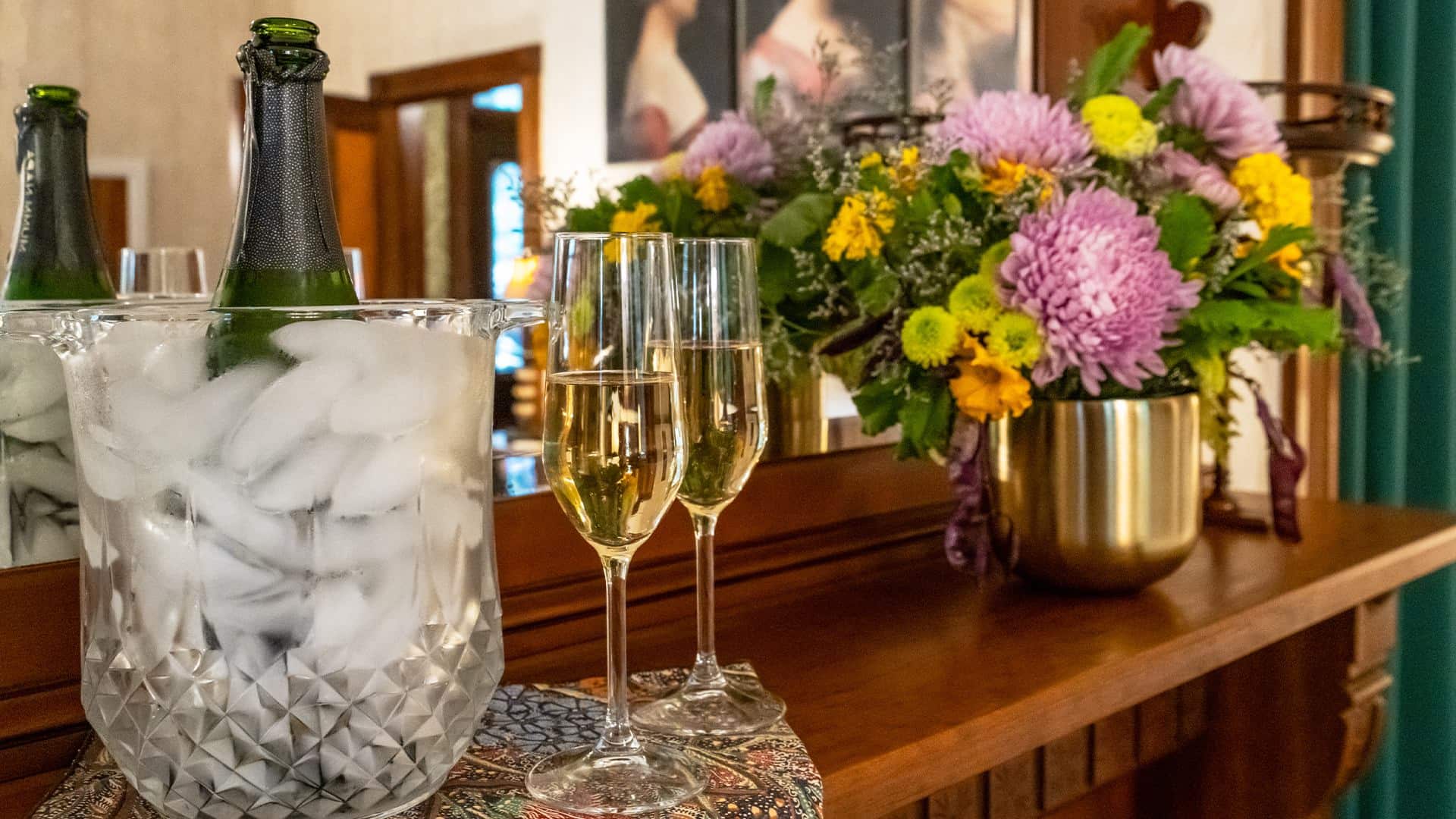 Close up view of a bottle chilling in a vase with ice, two glasses full of Champagne, and a vase of flowers on a wooden mantel
