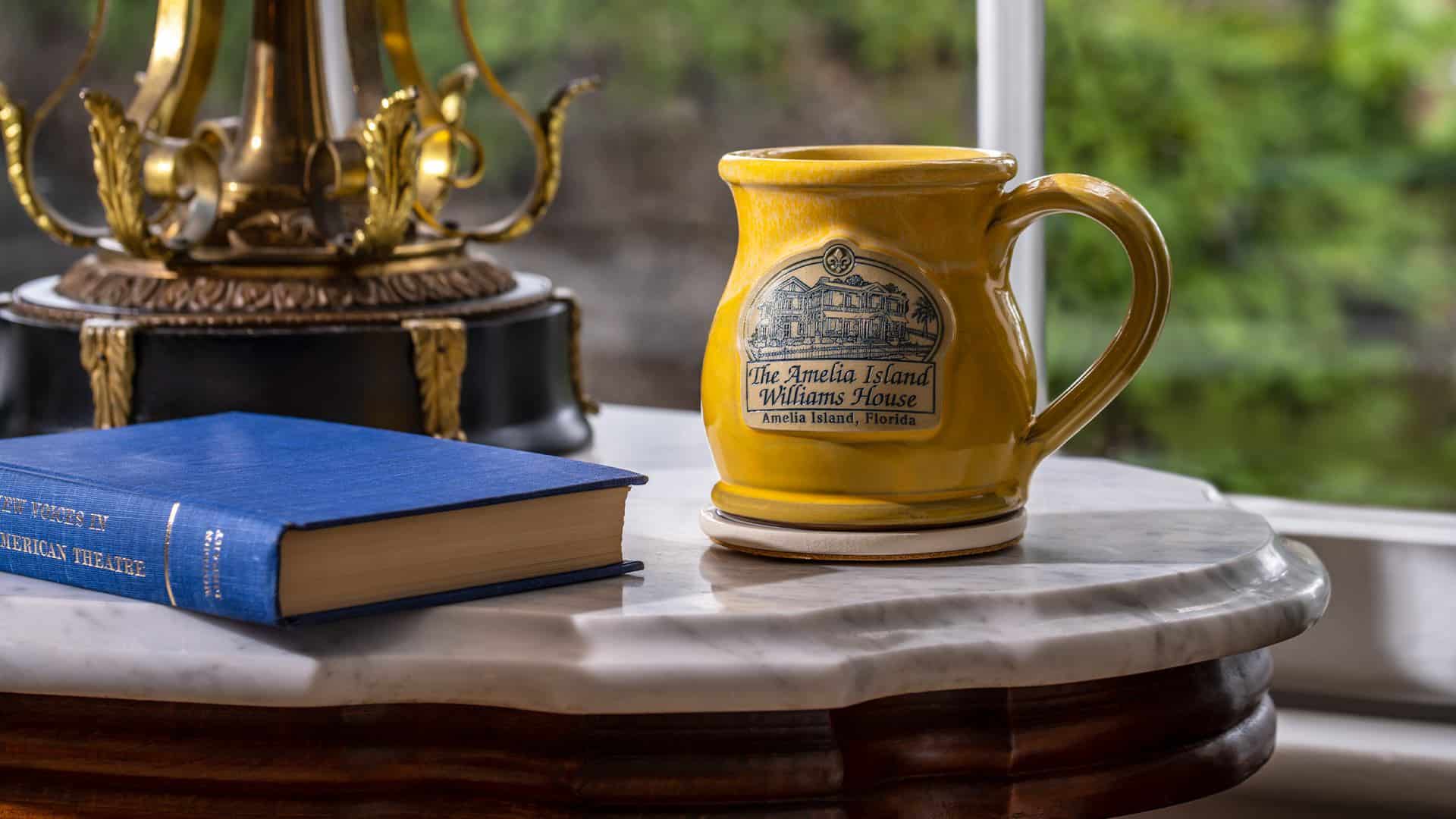 Close up view of yellow stoneware mug with The Amelia Island Williams House logo and blue book sitting on wooden stool with marble top