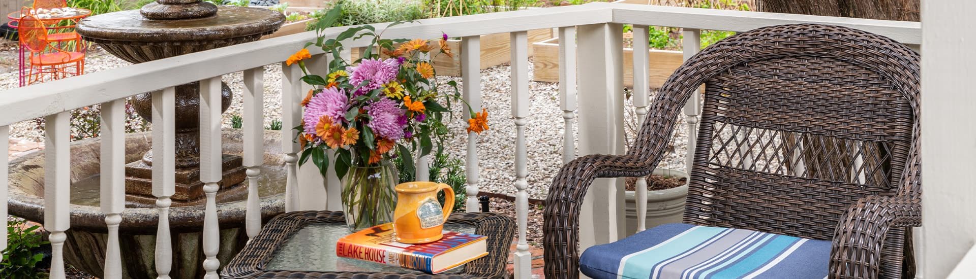 Close up view of purple and orange flowers in vase, orange stoneware mug, and book sitting on dark wicker patio table next to patio chair