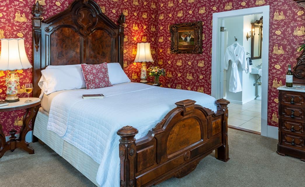 Bedroom with burgundy and gold ornate wallpaper, light-colored carpeting, dark wooden bed and dresser, white bedding, and view into bathroom