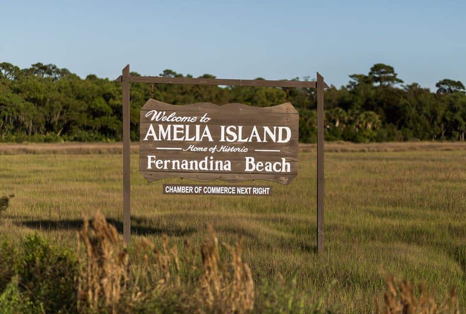 Wooden sign stating Welcome to Amelia Island in the middle of a grassy field with green trees in the background