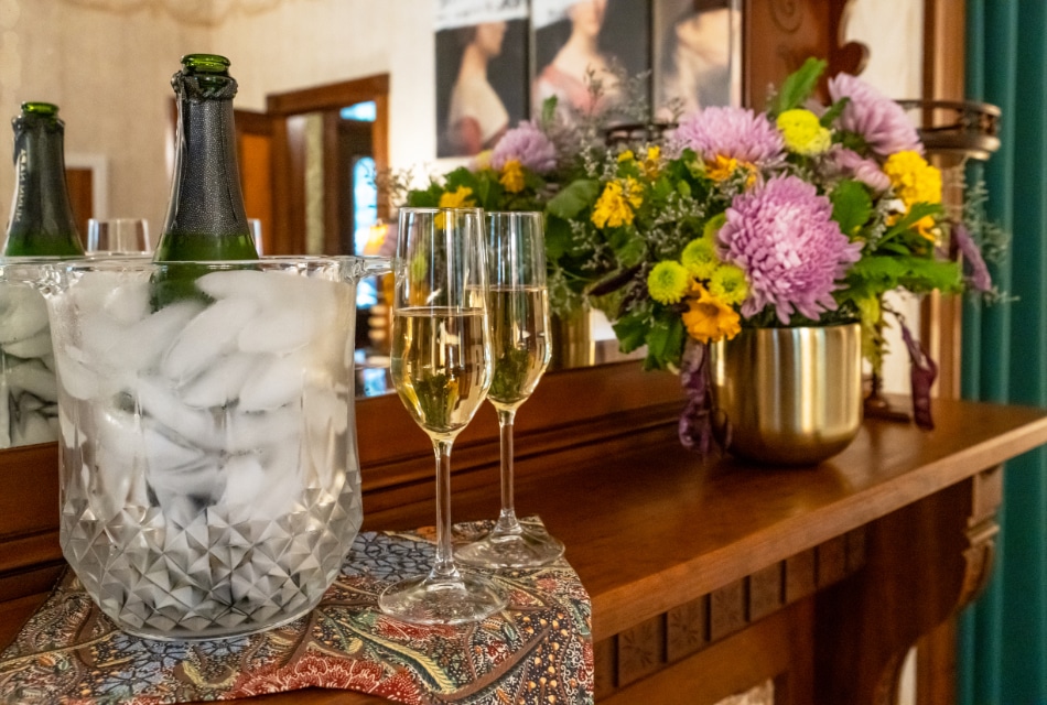 Close up view of a bottle chilling in a vase with ice, two glasses full of Champagne, and a vase of flowers on a wooden mantel
