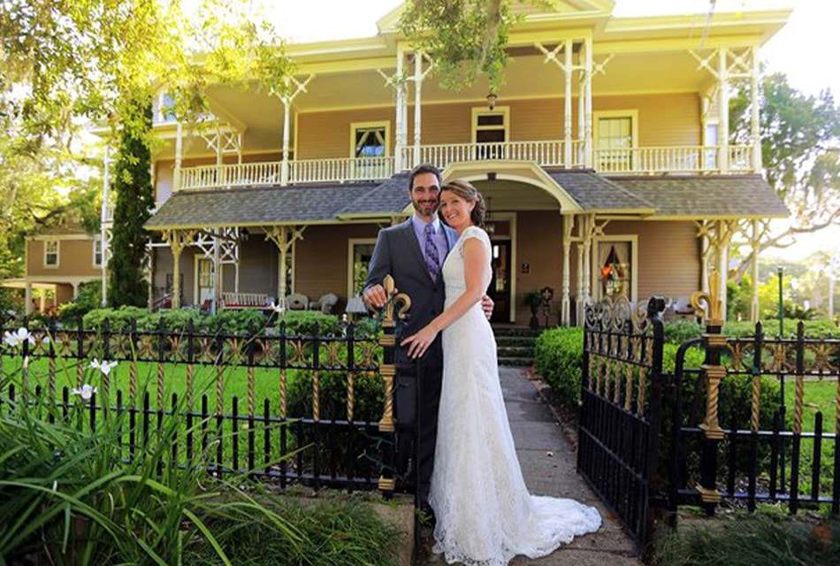 Woman in white dress and man in dark gray suit standing close to each other by wrought iron fence with property in the background